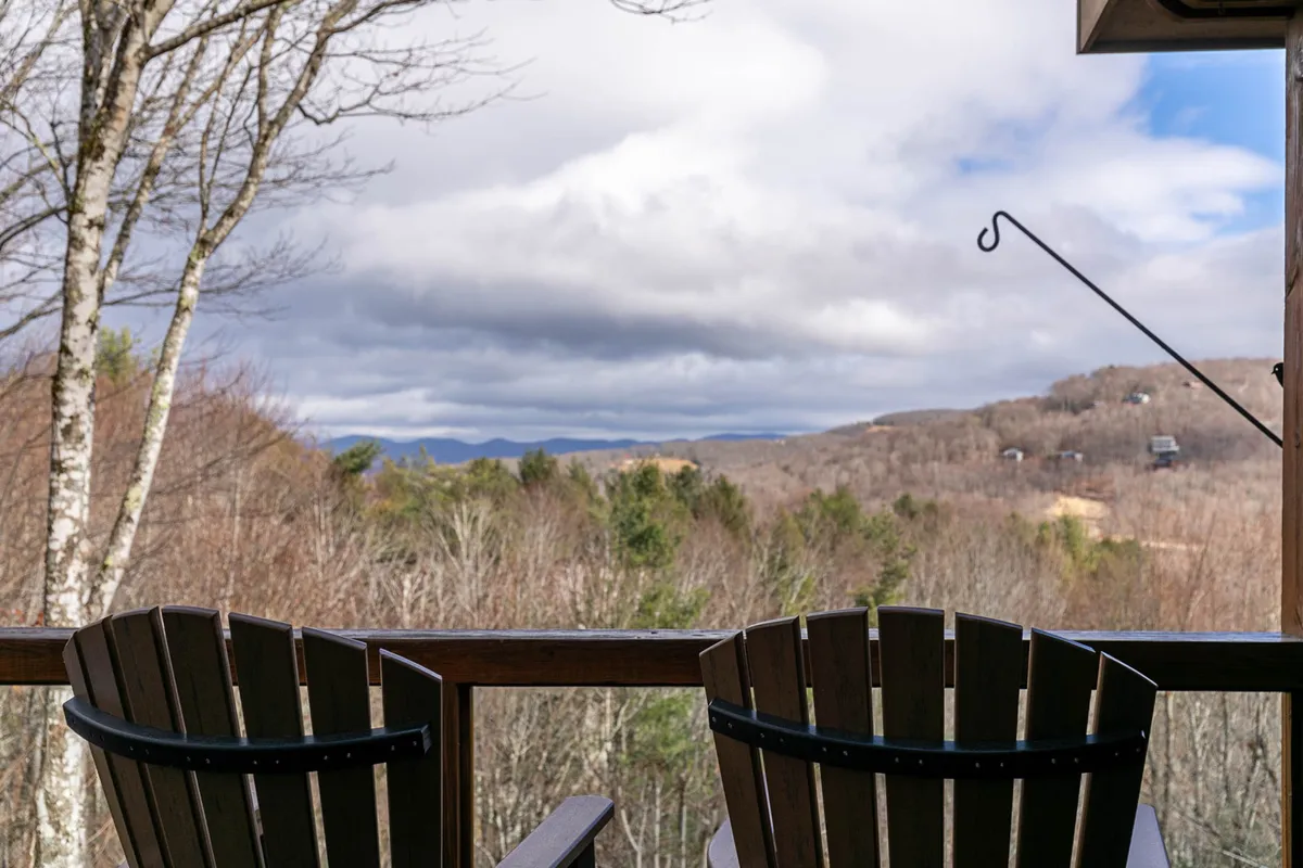 This image features two wooden chairs set on a deck or outdoor space, overlooking a peaceful mountain view beneath a cloudy sky. The natural landscape dominates the background, creating a serene and inviting atmosphere for relaxation and enjoying the outdoors with a pet.