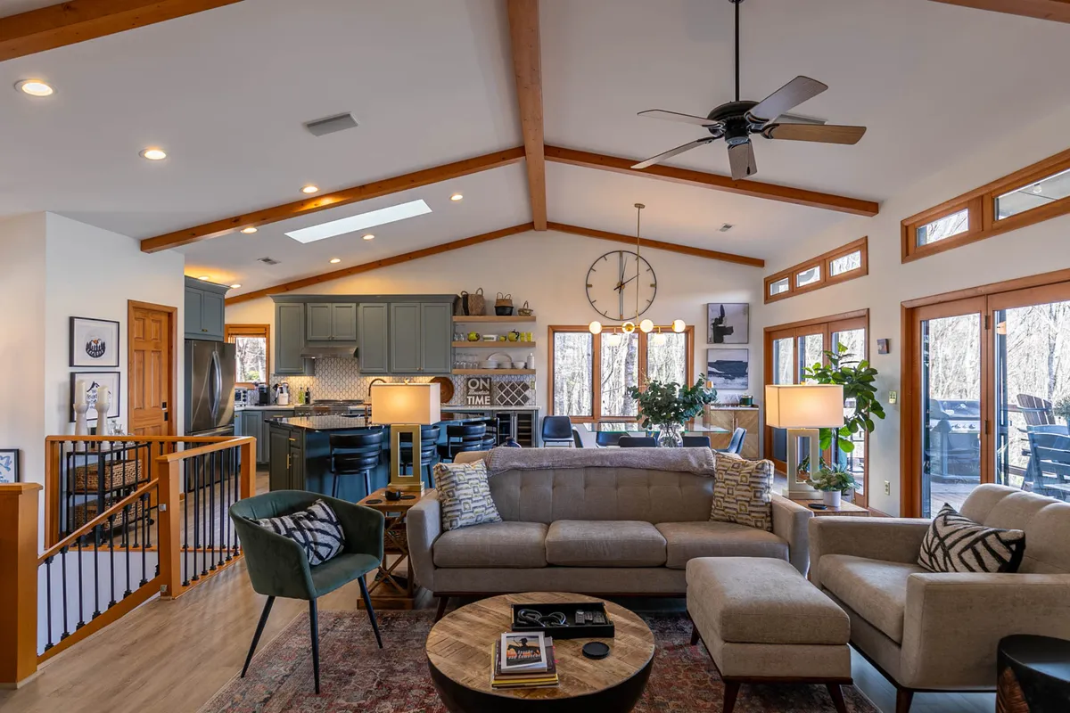 The image showcases a warm and inviting open-plan living area, featuring a plush sofa and coffee table perfect for gathering, a modern kitchen ready for shared meals, and expansive windows filling the space with natural light and offering glimpses of the mountain scenery outside.