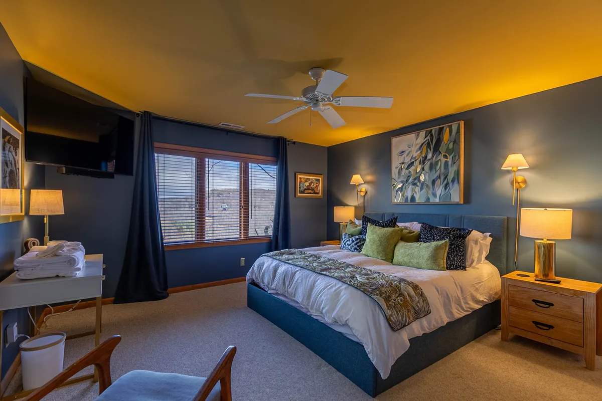 A beautifully made bed with decorative pillows sits beneath a large window fitted with blinds. Nightstands with lamps flank the bed, and calming artwork hangs on the wall. The serene setup offers a soothing, comfortable environment ideal for relaxation and unwinding after a day in the mountains.