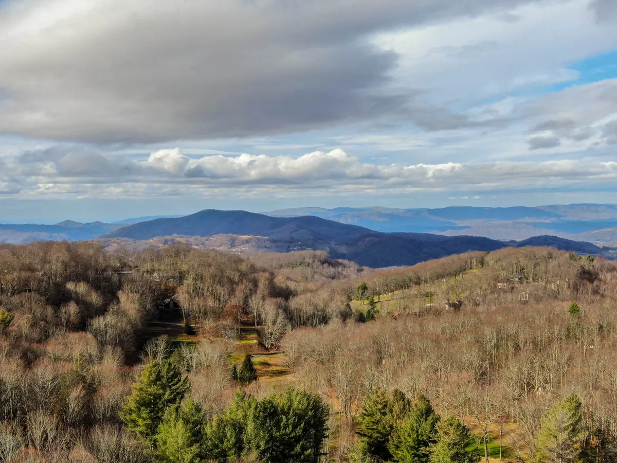 A breathtaking view of a mountain range at sunrise, with bare and evergreen trees in the foreground and a cloudy sky overhead. The scene is peaceful and expansive, reflecting the beauty and tranquility of Beech Mountain's natural landscape.