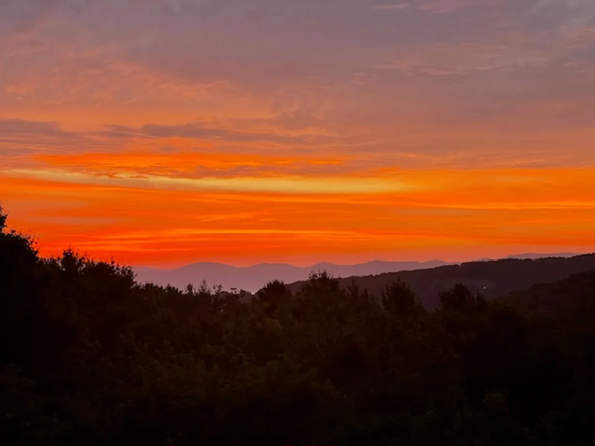 A stunning mountain sunset scene, with vibrant colors painting the sky above lush greenery and silhouetted peaks. The landscape invites viewers to imagine tranquil evenings outdoors.