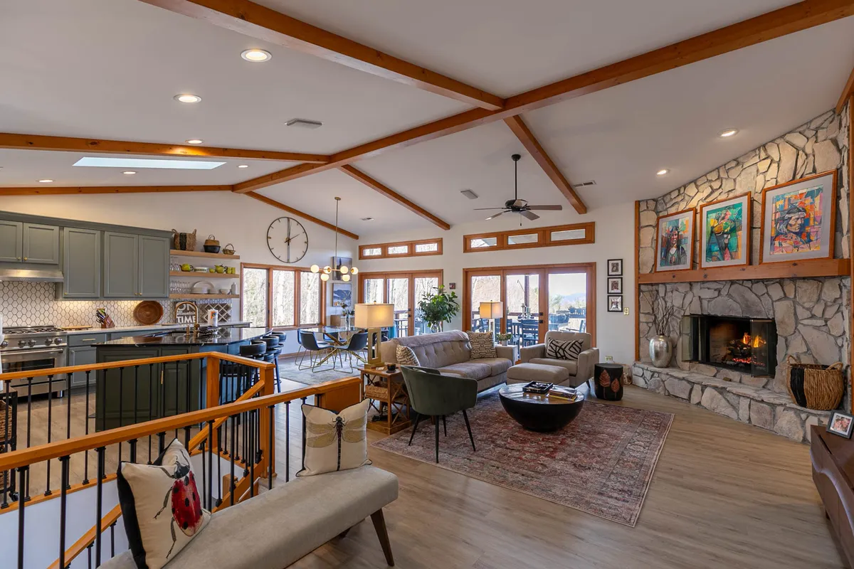 This image captures the heart of the communal living space, featuring a cozy fireplace as the focal point, a plush seating arrangement, a visible kitchen area, and large windows letting in natural light. The overall mood is welcoming and inviting.