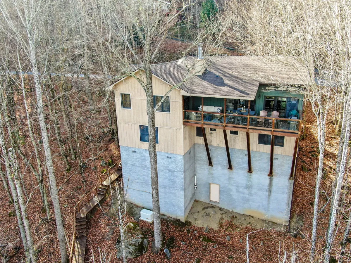 The photo captures the elevated exterior of The Raven, surrounded by mature trees and natural landscaping. A wooden deck extends from the home, and a visible pathway leads through the woods, suggesting immediate access to outdoor adventures and nearby trails. The setting is tranquil and embodies the spirit of exploration and family-friendly activities in nature.