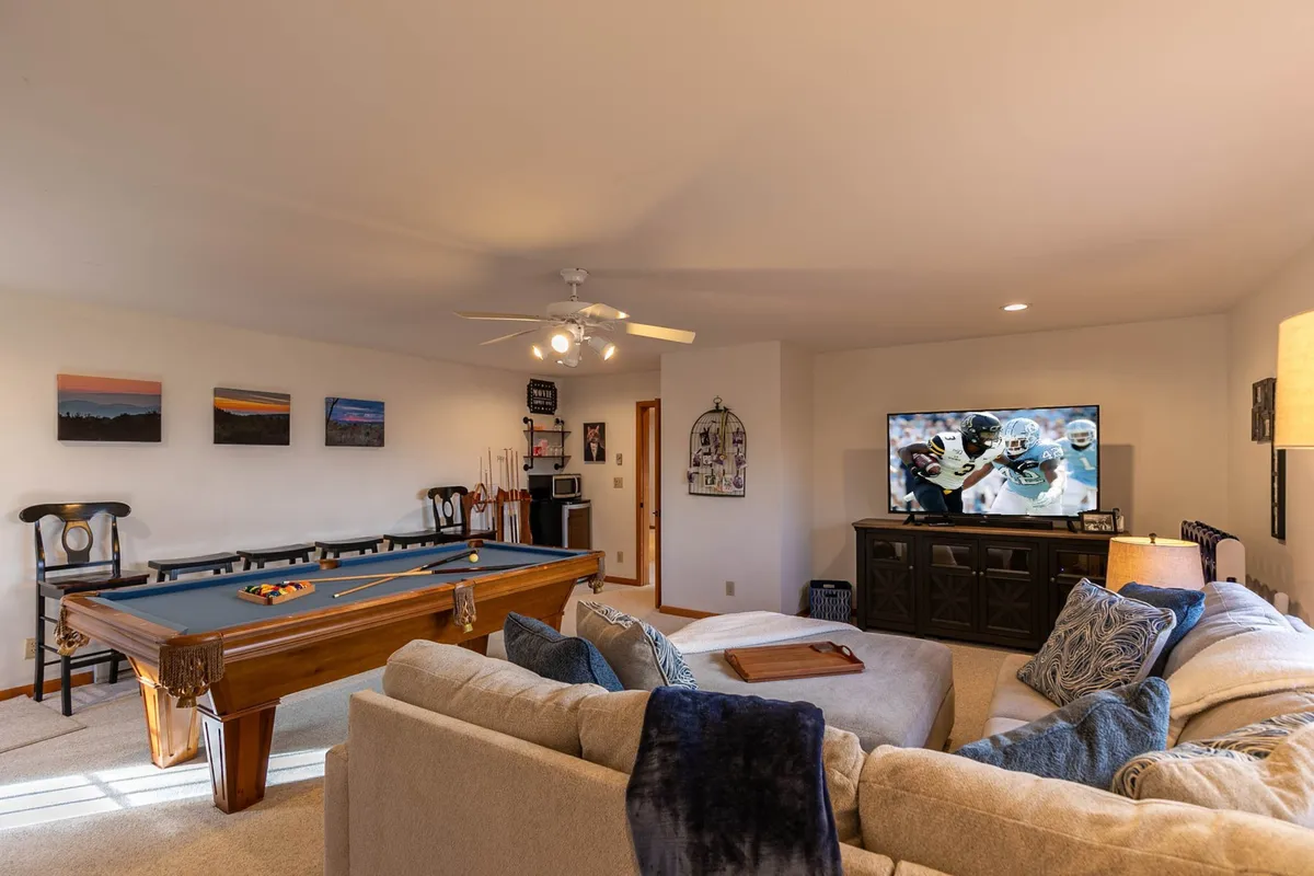A spacious, modern game room featuring a pool table, a large flatscreen TV for movie nights or sports viewing, a comfortable sofa, and vibrant wall art. The arrangement encourages both play and relaxation.